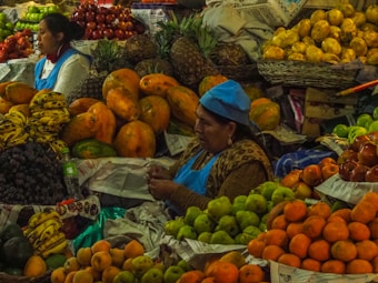 A bustling marketplace scene featuring two women in blue aprons surrounded by an abundance of colorful fruits. Various fruits are arranged in piles, including bananas, grapes, oranges, apples, and papayas. The women appear to be vendors, engaging in their daily routine amidst the vibrant display of produce.