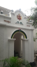 An ornate white archway featuring decorative elements like a crest and colorful accents. The arch is surrounded by greenery and has columns with decorative details. The numbers 1929 are inscribed, and text in a script is present. There is a pathway leading through the arch.