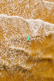 An aerial view of a single kayaker paddling through a vast expanse of foamy, golden-brown water. The waves create intricate patterns on the surface as they ripple around the kayak.