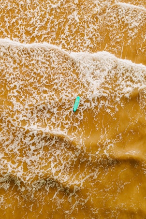 An aerial view of a single kayaker paddling through a vast expanse of foamy, golden-brown water. The waves create intricate patterns on the surface as they ripple around the kayak.