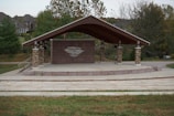 A small outdoor amphitheater with a wooden roof supported by stone pillars. The stage features a brick backdrop with a large emblem. Trees surround the structure, and residential homes are visible in the background.