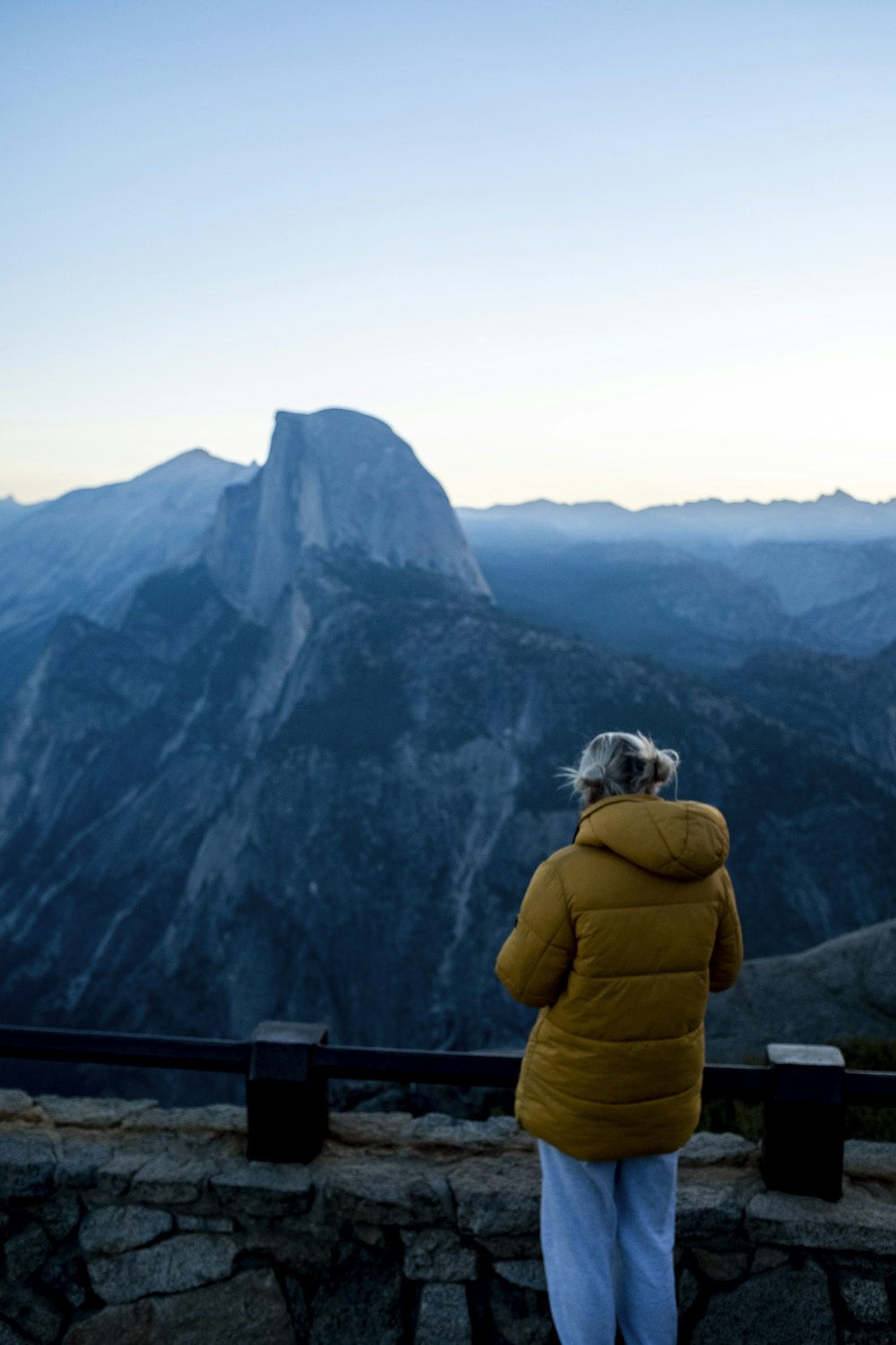 person in yellow jacket sitting on brown wooden fence looking at green mountains during daytime