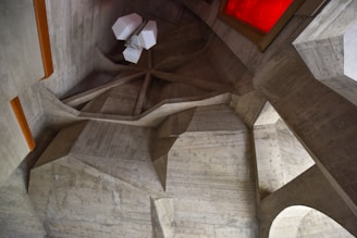 A geometric and abstract interior view featuring angular, concrete surfaces with sharp edges. The architecture presents a modern and industrial aesthetic. A unique, multi-faceted white light fixture hangs from the ceiling, adding contrast to the raw concrete texture. A bright red, rectangular element on the wall breaks the monochromatic scheme, injecting a splash of color.