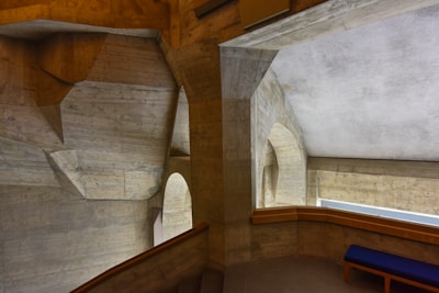 A concrete interior features angular, geometric structures with arches and linear patterns. The materials give the space an industrial yet modern aesthetic, complemented by muted lighting. A bench with a blue cushion is positioned against one wall, adding a touch of color.