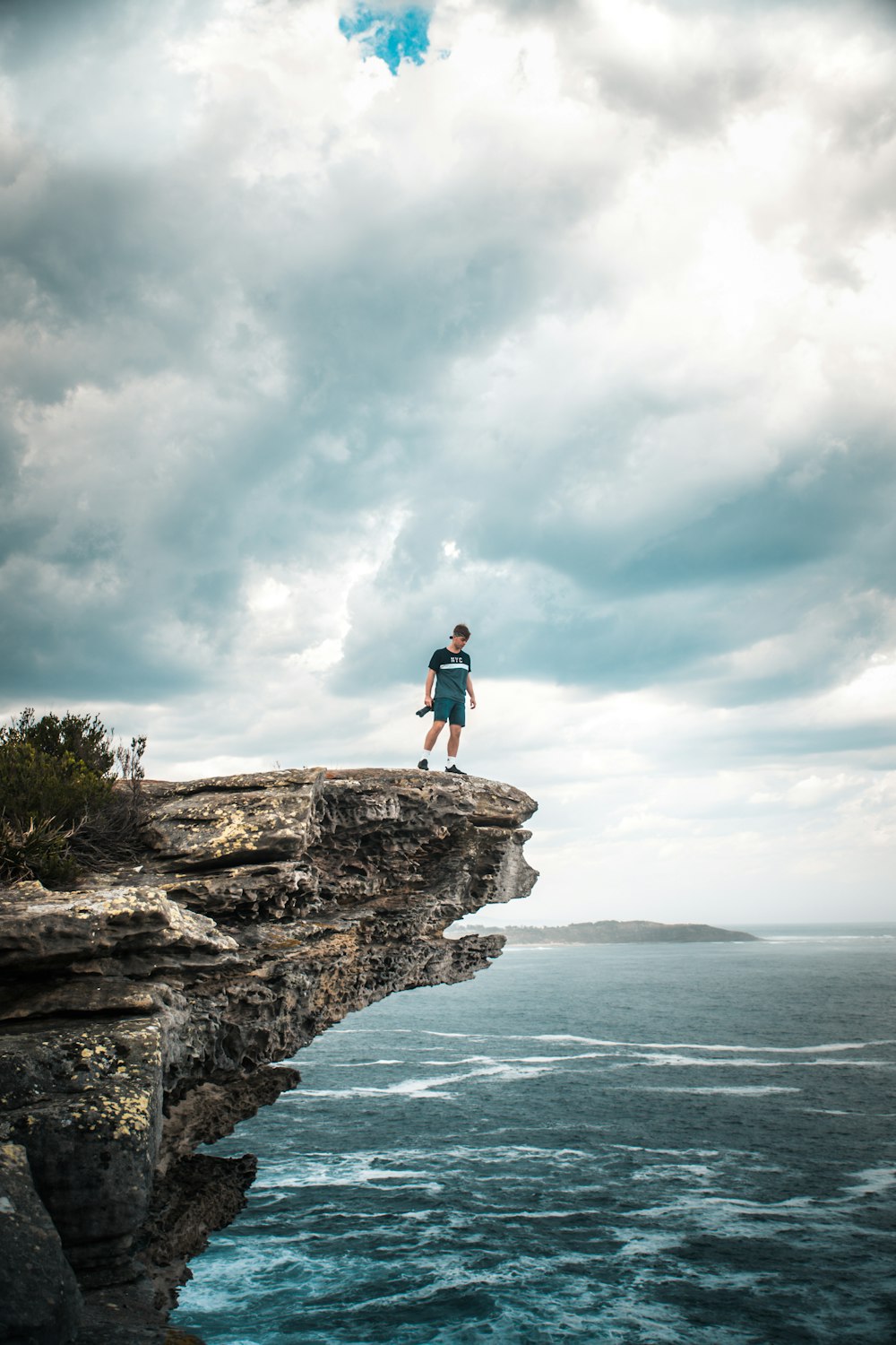 man in blue shirt and black shorts standing on rock formation near sea under white clouds