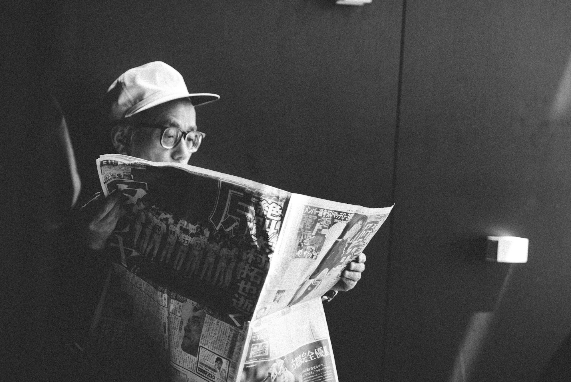 Black and white image of a man with a hat and glasses reading a newspaper