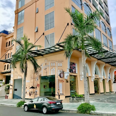A multistory building with a modern design features large windows and a decorative mural on the side wall. Two palm trees are in front, adding greenery to the urban setting. A parked car with a logo is visible on the street nearby.