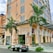 A multistory building with a modern design features large windows and a decorative mural on the side wall. Two palm trees are in front, adding greenery to the urban setting. A parked car with a logo is visible on the street nearby.