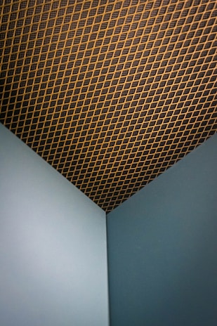 A corner of a room with two intersecting walls painted in a muted teal color. The ceiling has a grid-like pattern, featuring a mesh with gold or brass diagonal lines against a dark background.