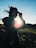 A silhouette of a person holding binoculars against the backdrop of a bright sun. The sunbeam creates a lens flare effect, while the person's hair is tousled by the wind. The scene appears to be outdoors, likely on a grassy hill or field with a clear blue sky above.