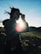 A silhouette of a person holding binoculars against the backdrop of a bright sun. The sunbeam creates a lens flare effect, while the person's hair is tousled by the wind. The scene appears to be outdoors, likely on a grassy hill or field with a clear blue sky above.