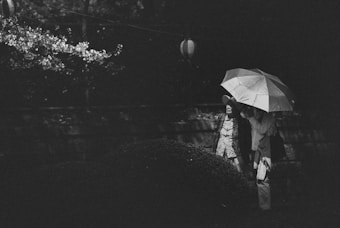 A black-and-white photograph capturing two people under an umbrella, standing near a bush and illuminated by a string of hanging lanterns. Their clothing suggests a casual outing, while the dim lighting adds a quiet, contemplative mood.