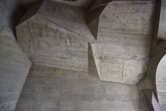 An interior ceiling made of concrete, featuring angular and geometric shapes with visible wood grain textures from the mold. The surface appears worn and has a grayish tone with variations due to shadows and lighting.