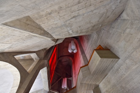 Concrete architectural structure with angular shapes, featuring a combination of sharp and geometric forms. A prominent red lighting section contrasts with the neutral tones of the concrete. Several modern light fixtures are hanging from the ceiling.