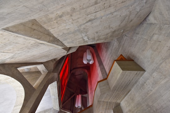 Concrete architectural structure with angular shapes, featuring a combination of sharp and geometric forms. A prominent red lighting section contrasts with the neutral tones of the concrete. Several modern light fixtures are hanging from the ceiling.
