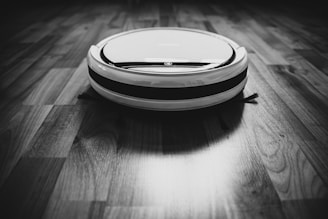 A robotic vacuum cleaner is situated on a wooden floor, captured in black and white. The device is circular with a sleek design, featuring contrasting stripes. The flooring displays a pattern of natural wood grain.
