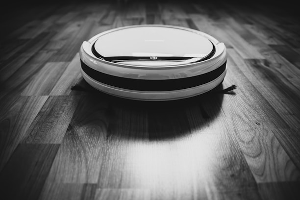 A robotic vacuum cleaner is situated on a wooden floor, captured in black and white. The device is circular with a sleek design, featuring contrasting stripes. The flooring displays a pattern of natural wood grain.
