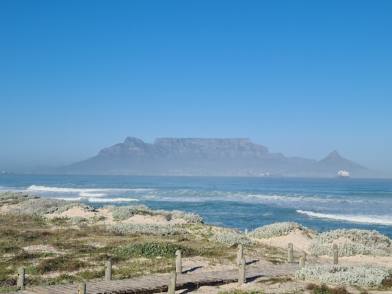 A scenic view of a beach with gentle waves lapping the shore, leading the eye to the majestic Table Mountain in the background. The sky is clear, allowing for a full view of the mountain's iconic flat top. A small ship is visible on the ocean to the right.