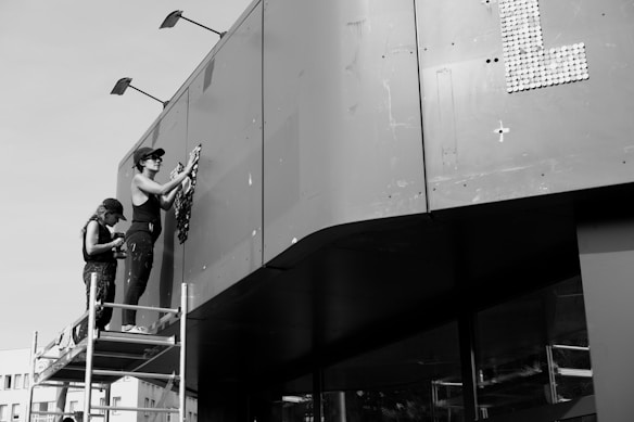 Two people are working on a large structure while standing on scaffolding. One person is applying materials to the surface, possibly for artistic or repair purposes, while the other stands behind, holding tools or equipment. The setting appears industrial, and the building surface is smooth and utilitarian, with some panels visible. The photo is in black and white, adding a stark, timeless quality.