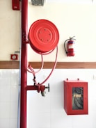 A red fire hose reel mounted on a wall alongside a fire extinguisher. The setup includes a red fire hose cabinet with a fire hose coiled inside, marked with the word 'FIRE' on the glass panel. The wall is tiled in white, contrasting with the red safety equipment.