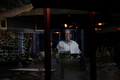 A person wearing a white chef's jacket is standing behind a glass wall with reflections of interior lights and objects such as bottles and a bowl visible. The scene appears to be inside a restaurant or a similar establishment, with a dark and somewhat cozy atmosphere.