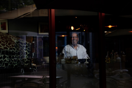 A person wearing a white chef's jacket is standing behind a glass wall with reflections of interior lights and objects such as bottles and a bowl visible. The scene appears to be inside a restaurant or a similar establishment, with a dark and somewhat cozy atmosphere.