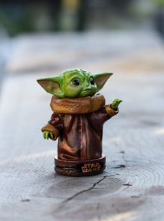 A small, detailed figurine of a character with green skin, large ears, and oversized eyes, resembling a baby alien. The figure is wearing a brown robe with a high collar and is positioned on a wooden surface. It stands on a round base with the words 'Star Wars' inscribed on it.