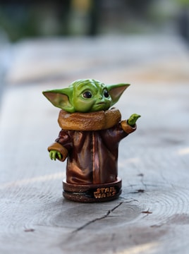 A small, detailed figurine of a character with green skin, large ears, and oversized eyes, resembling a baby alien. The figure is wearing a brown robe with a high collar and is positioned on a wooden surface. It stands on a round base with the words 'Star Wars' inscribed on it.