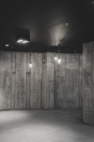 A minimalist interior featuring raw concrete walls and a smooth floor. The lighting is subdued, with a few hanging bulbs providing direct illumination and casting soft shadows across the surfaces.