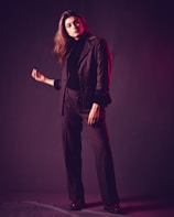 A person stands confidently with one hand raised slightly while dressed in a dark tailored suit. The lighting creates a soft, moody atmosphere, with a hint of pinkish-red hues reflecting on the suit.