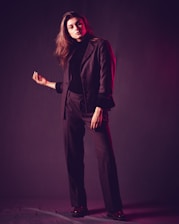 A person stands confidently with one hand raised slightly while dressed in a dark tailored suit. The lighting creates a soft, moody atmosphere, with a hint of pinkish-red hues reflecting on the suit.