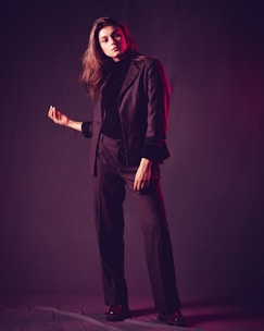 A person stands confidently with one hand raised slightly while dressed in a dark tailored suit. The lighting creates a soft, moody atmosphere, with a hint of pinkish-red hues reflecting on the suit.