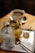 A tray holds a glass of orange slices and mint leaves mixed with ice, a small bottle of water, a glass shot with amber liquid likely tea or syrup, and a gold spoon. In the background, there is a cup of coffee sitting on a saucer, all placed on a wooden surface.