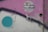 An abstract piece of street art featuring a mix of colors and shapes. The artwork includes a large turquoise circle, a zigzag arrow, and a word 'STREET ART' written in white within a circle. The background blends shades of purple, grey, and beige with textured and weathered surfaces.