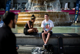 A couple laughing together as they read clues next to a historic fountain in a sunny town square.