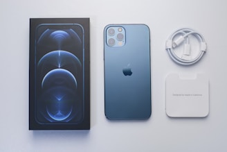 A smartphone with a blue finish placed next to its packaging box, a white charging cable, and a white manual packet. The phone features a sleek design with multiple camera lenses on the back.