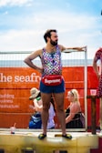 A person stands confidently on a raised platform, dressed in a colorful, sleeveless, scale-patterned top and dark denim shorts. They wear a red pouch with a prominent brand logo around their waist and black slip-on shoes with a yellow emblem. The background shows a bright orange structure with partially visible text and faint outlines of other people and objects.