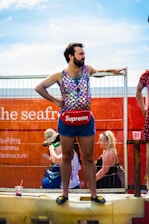 A person stands confidently on a raised platform, dressed in a colorful, sleeveless, scale-patterned top and dark denim shorts. They wear a red pouch with a prominent brand logo around their waist and black slip-on shoes with a yellow emblem. The background shows a bright orange structure with partially visible text and faint outlines of other people and objects.