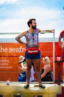 A person stands confidently on a raised platform, dressed in a colorful, sleeveless, scale-patterned top and dark denim shorts. They wear a red pouch with a prominent brand logo around their waist and black slip-on shoes with a yellow emblem. The background shows a bright orange structure with partially visible text and faint outlines of other people and objects.