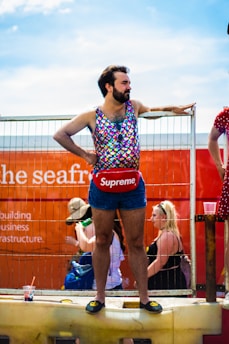 A person stands confidently on a raised platform, dressed in a colorful, sleeveless, scale-patterned top and dark denim shorts. They wear a red pouch with a prominent brand logo around their waist and black slip-on shoes with a yellow emblem. The background shows a bright orange structure with partially visible text and faint outlines of other people and objects.