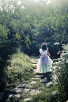 A child dressed in a whimsical costume with wings and a pastel-colored tulle skirt walks along a stone path surrounded by lush greenery and white flowers, bathed in soft sunlight.