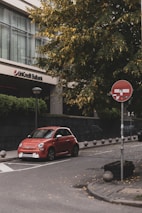 A red compact car is parked on a city street near a tall building with the sign 'UniCredit Bulbank'. The street is lined with stone bollards and has a stop sign with a graphic. Green trees with yellowing leaves are visible in the background, adding a touch of nature to the urban setting.