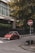 A red compact car is parked on a city street near a tall building with the sign 'UniCredit Bulbank'. The street is lined with stone bollards and has a stop sign with a graphic. Green trees with yellowing leaves are visible in the background, adding a touch of nature to the urban setting.