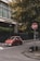A red compact car is parked on a city street near a tall building with the sign 'UniCredit Bulbank'. The street is lined with stone bollards and has a stop sign with a graphic. Green trees with yellowing leaves are visible in the background, adding a touch of nature to the urban setting.