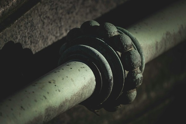 A close-up view of a metal pipe with a decorative, rounded metal element that seems to be part of an industrial or architectural structure. The surface shows signs of aging with spots and a patina, suggesting weathered material. The image is shadowy, with strong contrasts between light and dark areas.