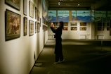 An art gallery with several framed paintings displayed on the walls. A person stands in front of the artwork, seemingly taking pictures or observing closely. The lighting is soft and focused on the paintings, creating a serene and contemplative atmosphere.