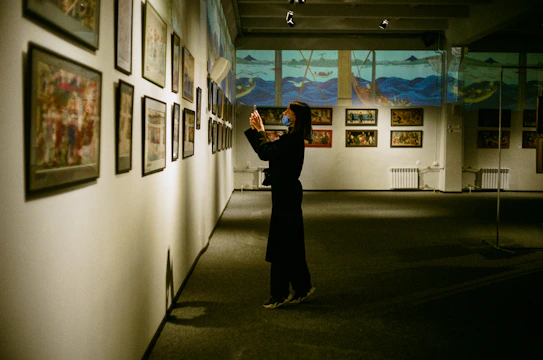 An art gallery with several framed paintings displayed on the walls. A person stands in front of the artwork, seemingly taking pictures or observing closely. The lighting is soft and focused on the paintings, creating a serene and contemplative atmosphere.