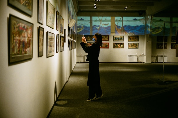 An art gallery with several framed paintings displayed on the walls. A person stands in front of the artwork, seemingly taking pictures or observing closely. The lighting is soft and focused on the paintings, creating a serene and contemplative atmosphere.