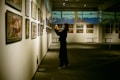 An art gallery with several framed paintings displayed on the walls. A person stands in front of the artwork, seemingly taking pictures or observing closely. The lighting is soft and focused on the paintings, creating a serene and contemplative atmosphere.