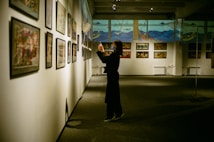 An art gallery with several framed paintings displayed on the walls. A person stands in front of the artwork, seemingly taking pictures or observing closely. The lighting is soft and focused on the paintings, creating a serene and contemplative atmosphere.